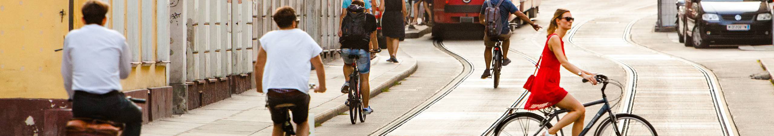 Mehrere Personen fahren mit Fahrrädern entlang der Straße, im Vordergrund eine alte Wiener Straßenbahn. Eine Frau in einem roten Sommerkleid auf einem Hollandrad quert die Fahrbahn. Foto von Peter Provaznik