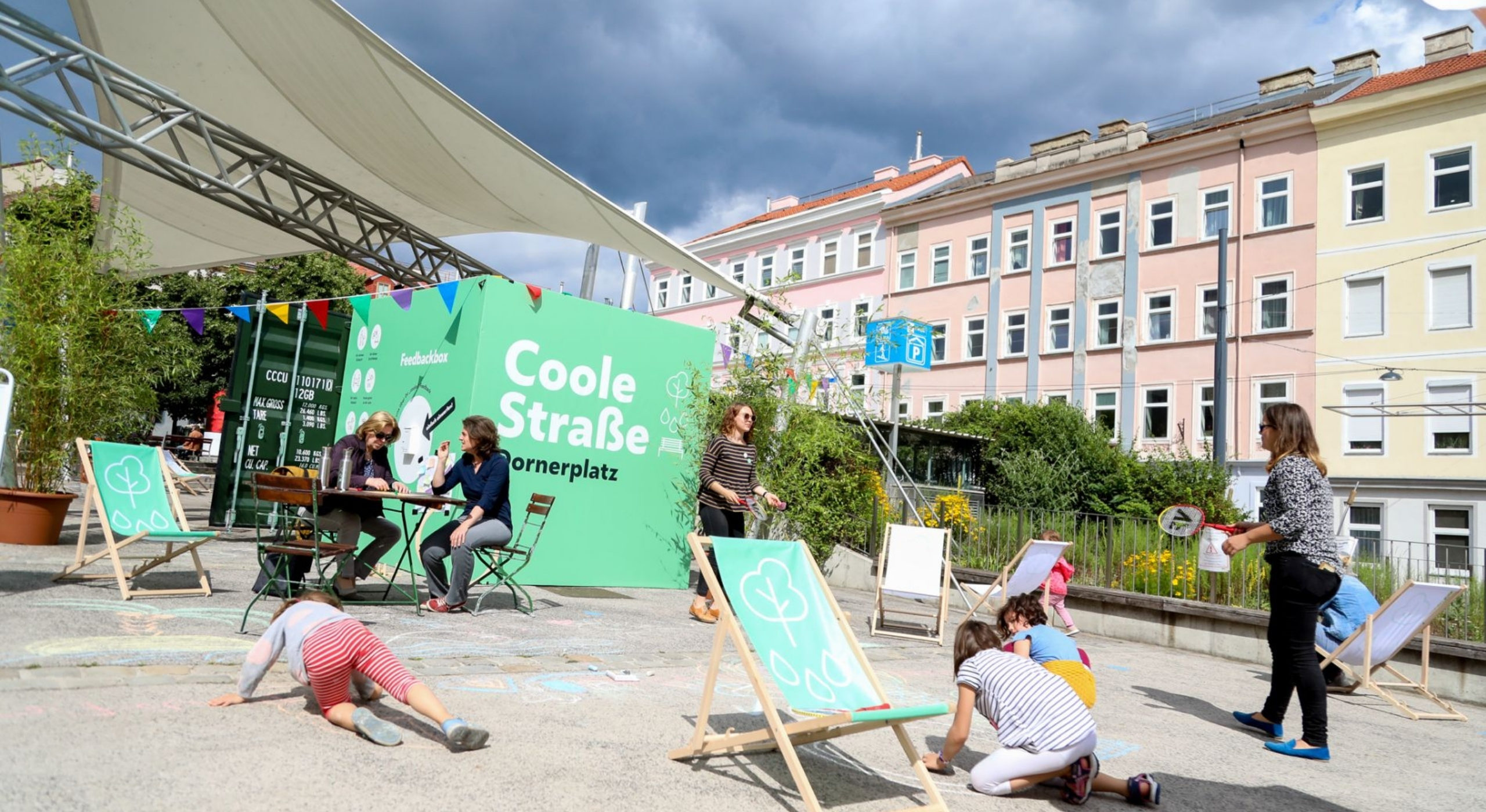 Foto der Coolen Straße. Kinder bemalen die Straße mit Kreide, spielen Federball. Zwei Frauen sitzen im Schatten und unterhalten sich.