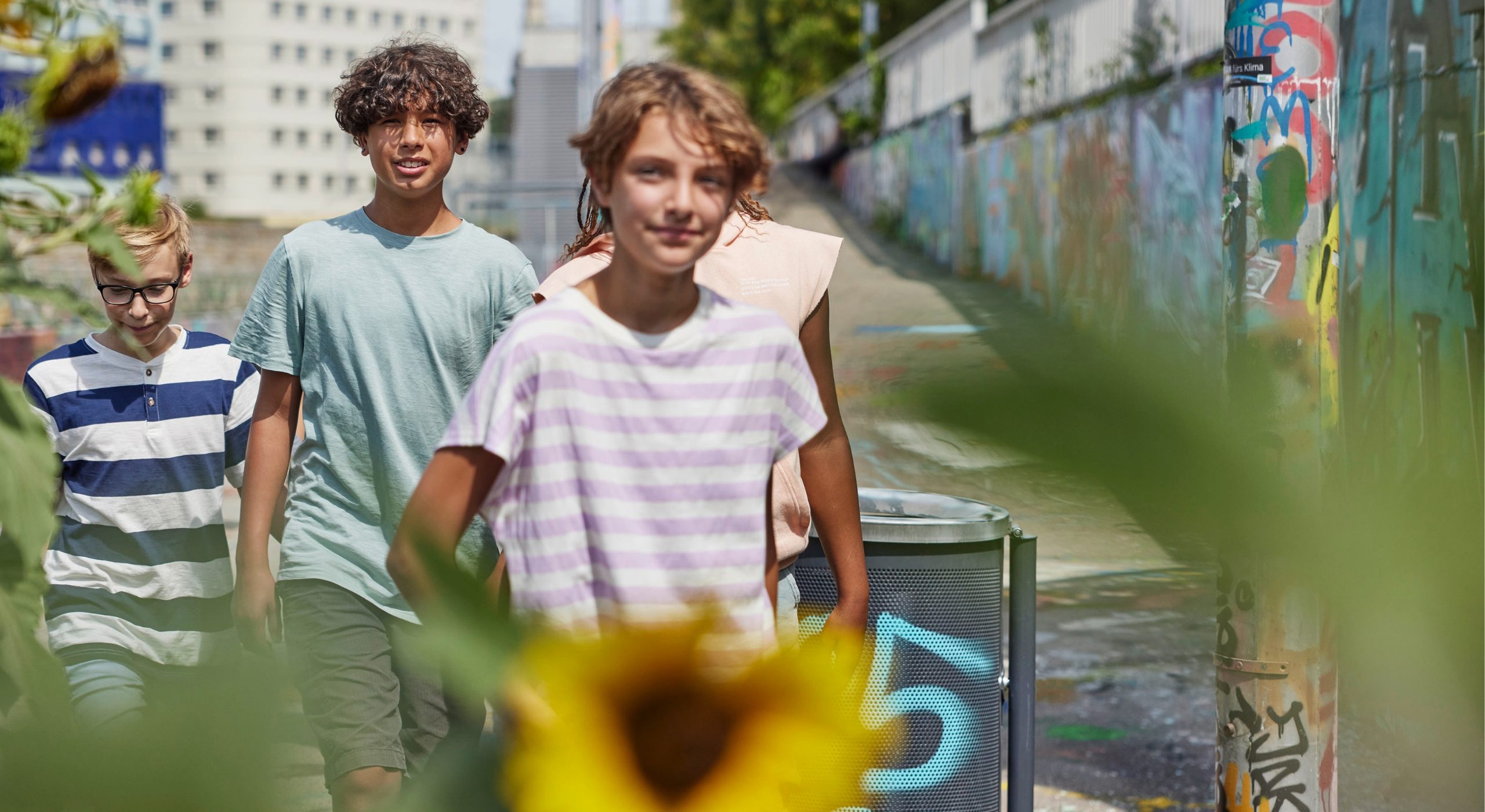 Drei junge Leute, sie sind etwa 13 Jahre alt, gehen gemeinsam spazieren. Im Vordergrund sieht man eine Sonnenblume.