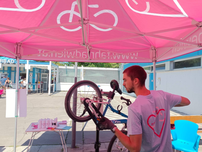 Mechaniker repariert ein Fahrrad. Im Hintergrund sieht man den Eingang zu einem Svhwimmbad.